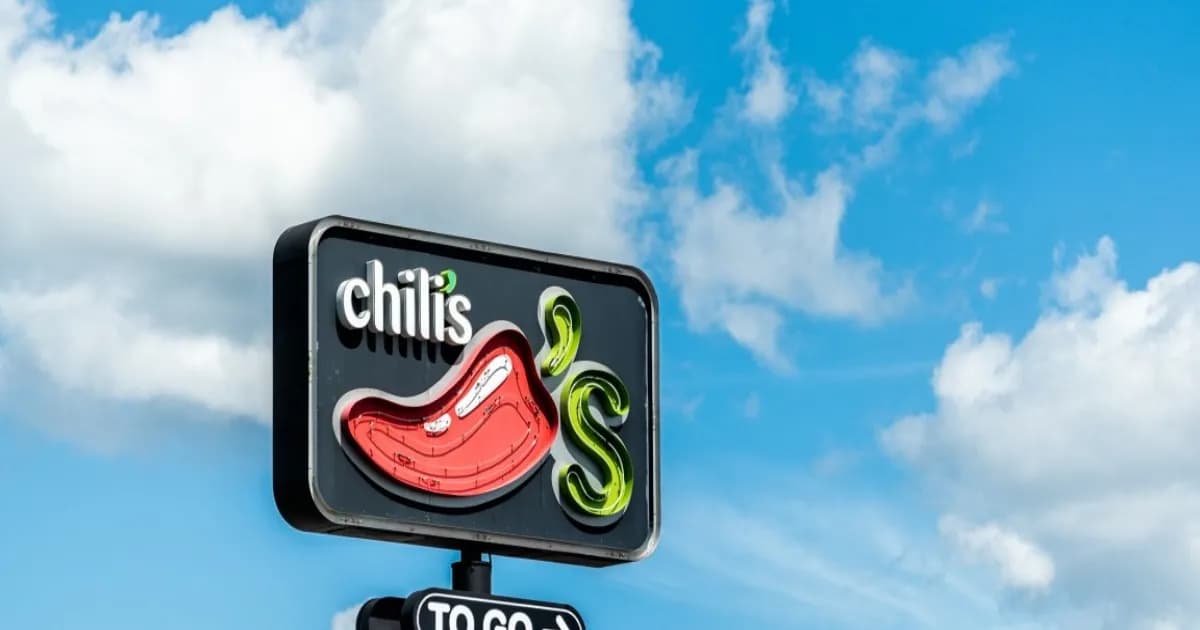 Papan nama restoran Chili's dengan logo cabai merah dan teks "Chili's" di latar belakang hitam, dengan papan tambahan "TO GO" di bawahnya, berlatar langit biru dengan awan putih.